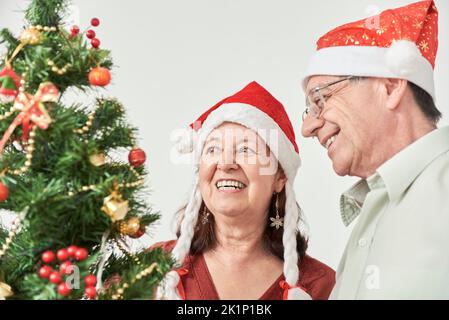 Joyeux couple hispanique senior souriant tout en décorant leur arbre de Noël, portant des béanies rouges du Père Noël. La joie de passer les vacances ensemble Banque D'Images