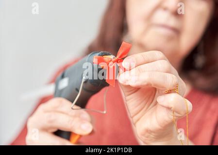 Femme adulte non reconnaissable faisant des métiers de Noël, mains utilisant un pistolet en silicone chaud pour coller un arc rouge. Banque D'Images