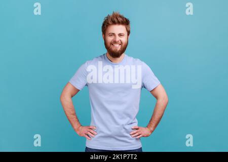 Portrait d'un homme à barbe heureux et satisfait debout avec les mains sur les hanches et regardant l'appareil photo avec un sourire en dents de souris, ayant une expression positive et confiante. Studio d'intérieur isolé sur fond bleu. Banque D'Images