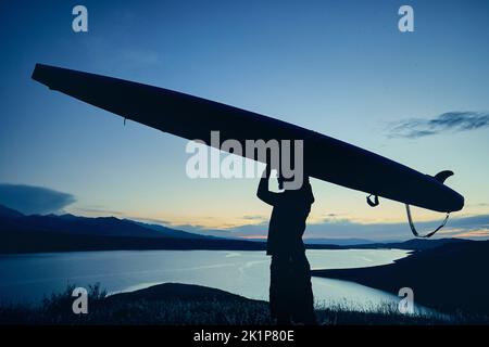 Homme barbu en silhouette tenant le panneau SUP près du lac au coucher du soleil au Kazakhstan. Levez-vous paddle board en plein air, loisirs actifs dans la nature. Banque D'Images