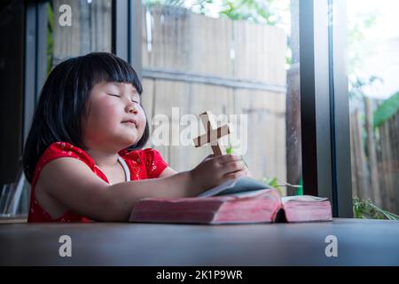 Little girl praying and holding wooden cross on bible at home, child's pure faith, focus at face. Stock Photo