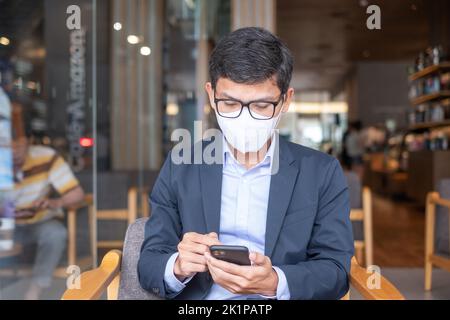 Jeune homme d'affaires en costume portant un masque chirurgical et en utilisant un smartphone, homme dactylographiant l'écran tactile téléphone mobile dans le bureau ou le café. Pandémie Covid-19, t Banque D'Images