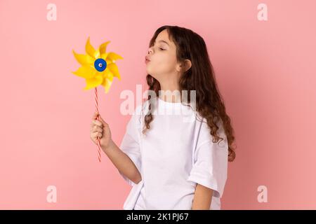 Portrait de charmante petite fille amusante et adorable portant un T-shirt blanc soufflant au moulin à vent en papier, jouant avec un jouet à volant sur bâton. Studio d'intérieur isolé sur fond rose. Banque D'Images