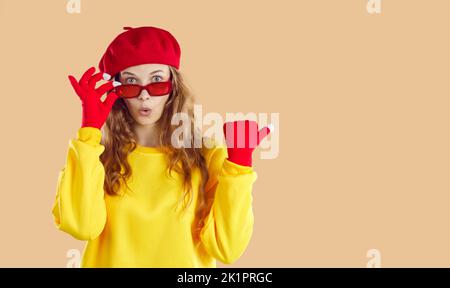 Femme surprise en sweat-shirt jaune, chapeau rouge, gants lunettes regardant la caméra sur fond beige. Banque D'Images