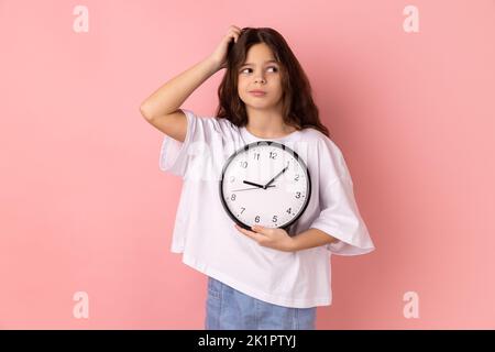 Portrait d'une petite fille perplexe portant un T-shirt blanc tenant une horloge murale et se grattant la tête, échéance, ponctualité. Studio d'intérieur isolé sur fond rose. Banque D'Images