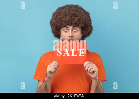 Homme avec la coiffure d'Afro portant un T-shirt orange debout avec le mot de vente, regardant avec les grands yeux surpris, choqué par les réductions, Black Friday bas prix. Studio d'intérieur isolé sur fond bleu. Banque D'Images