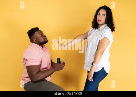 Portrait d'un jeune couple dans des vêtements décontractés, homme faisant une proposition à sa petite amie, femme refusant, montrant stop geste de paume. Studio d'intérieur isolé sur fond jaune. Banque D'Images