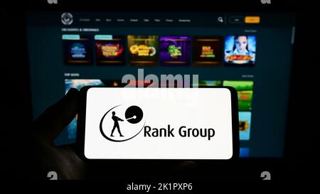 Personne tenant un smartphone avec le logo de la compagnie de jeu britannique The Rank Group plc à l'écran devant le site Web. Mise au point sur l'affichage du téléphone. Banque D'Images
