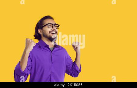 Un jeune homme sud-asiatique souriant lève les mains comme un signe réussi, debout dans un studio jaune Banque D'Images