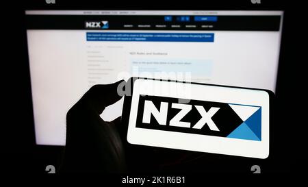 Personne tenant un téléphone portable avec le logo de la société financière New Zealand's Exchange (NZX) à l'écran en face de la page Web. Mise au point sur l'affichage du téléphone. Banque D'Images