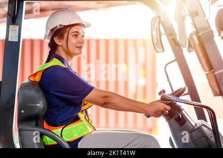 opérateur de chariot élévateur féminin travaillant dans un entrepôt. Portrait d'une jeune femme indienne chauffeur assis dans un chariot élévateur et souriant travaillant dans un grand entrepôt Banque D'Images