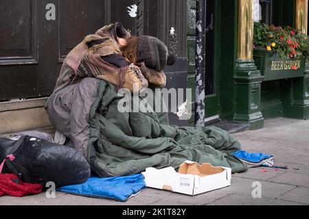 L'homme ou le bum sans-abri, enveloppé dans des couvertures et des sacs de couchage, s'assoit avec sa tête couverte sur le trottoir dans une rue commerçante à côté d'une boîte vide Banque D'Images