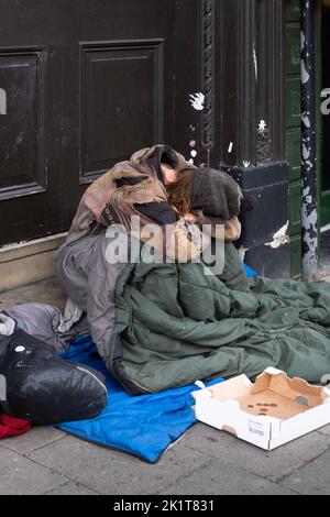 L'homme ou le bum sans-abri, enveloppé dans des couvertures et des sacs de couchage, s'assoit avec sa tête couverte sur le trottoir dans une rue commerçante à côté d'une boîte vide Banque D'Images