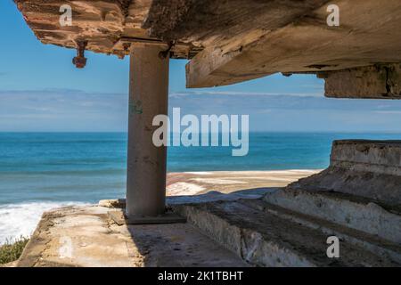 Un bunker de la Batterie des Arros au nord de Soulac-sur-Mer dans le Médoc, en France, possède un toit en béton massif qui dépasse largement, soutenu par une épaisse colonne en béton. La vue passe entre la colonne et le toit sur la plage de sable et l’Atlantique. Le site fait partie des derniers survivants d’environ 350 bunkers constituant la « Festung Gironde-Süd » (forteresse de Gironde Sud) près de Bordeaux, agrandie par la Wehrmacht allemande en 1943 sur une position de la Marine française datant de 1938 – une partie du mur de l’Atlantique. Des barres de renforcement rouillées dépassent du béton fissuré du plafond. Banque D'Images