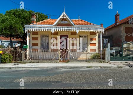 La villa „Obdélia“ se dresse rue Joseph Lahens à Soulac-sur-Mer, Médoc, France, son nom est indiqué sur une plaque ovale encastrée dans le pignon ornemental blanc. Une frise délicate en bois festonnée (lambrequin), peinte en blanc, court sous l’avant-toit au-dessus d’une porte d’entrée rouge encadrée de deux volets blancs patinés. Une clôture en grillage avec une porte en fer forgé et une boîte aux lettres verte se trouvent dans le jardin avant. La maison est l’une des nombreuses villas construites dans le style local „villa soulacaise“, prisé surtout par les familles aisées de Bordeaux après l’ouverture de la ligne ferroviaire Bordeaux–Soulac en 1874 – caractérisé par le contr Banque D'Images