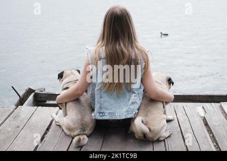 Jeune femme assise embrassant deux pugs près de la surface de l'eau. Style de vie avec des chiens, fille assise près de la rivière avec ses animaux de compagnie Banque D'Images