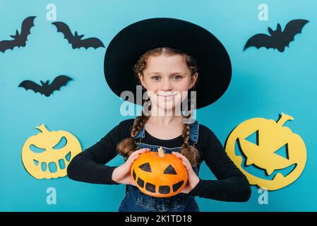 Joyeux Halloween et concept de vacances d'automne. Portrait de charmante petite fille dans un grand chapeau noir tenant une petite citrouille d'orange drôle, posant isolat Banque D'Images