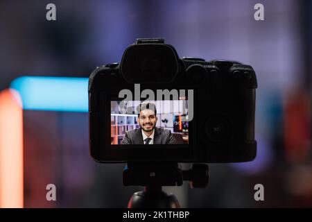 Vue depuis l'écran de la caméra de l'attrayant positif confiant expérimenté intelligent indien barbu bureau Manager enregistrement vidéo vlog pour le public Internet dans le bureau de soirée. Banque D'Images