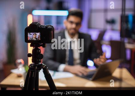 Vue depuis l'écran de la caméra de l'attrayant positif confiant expérimenté intelligent indien barbu bureau Manager enregistrement vidéo vlog pour le public Internet dans le bureau de soirée. Banque D'Images