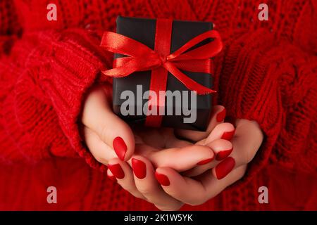 Mains de femmes manucures dans un chandail rouge contenant une boîte cadeau. Solde de Noël pour les Saint-Valentin Banque D'Images