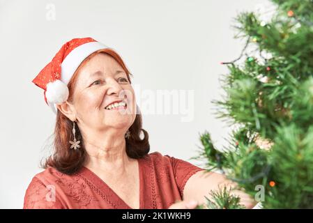 Heureuse femme hispanique senior souriant tout en décorant un arbre de Noël portant un chapeau rouge du Père Noël. La joie des vacances. Banque D'Images