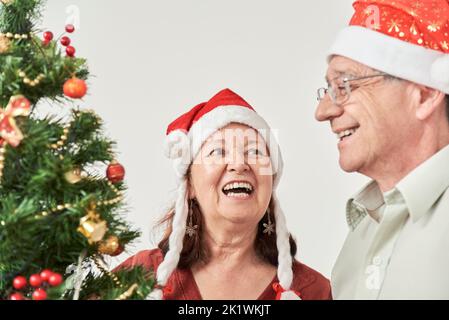 Joyeux couple hispanique senior souriant tout en décorant leur arbre de Noël, portant des béanies rouges du Père Noël. La joie de passer les vacances ensemble Banque D'Images