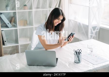 Travail à distance. Déménagement de bureaux. Ferme-tout. Une femme heureuse et inspirée travaillant à domicile sur un projet de démarrage avec un téléphone portable dans un nouvel éclairage A. Banque D'Images