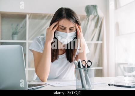 Ferme-tout. Déménagement au bureau. Le chômage de quarantaine. Stressé femme impatiente dans un masque facial de protection souffrant de maux de tête à la lumière Banque D'Images