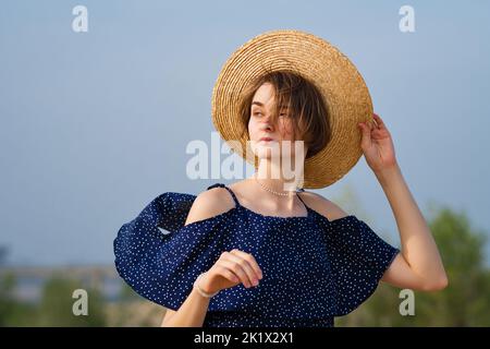 Gros plan portrait de la jeune femme caucasienne avec une coupe de cheveux courte. Fille porte un chapeau de paille et une robe bleu foncé avec des pois. La femelle regarde loin poser la nature Banque D'Images