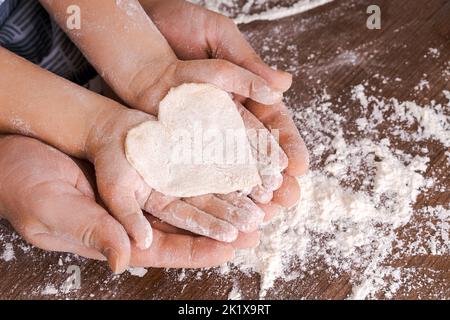 Vue de dessus la main du père et de l'enfant, découpez des biscuits en forme de coeur de la pâte sur les mains sur le fond d'une table en bois avec de la farine en vrac. Défaillance de l'écran plat. Banque D'Images