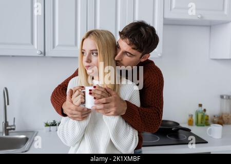 jeune homme en chandail rouge embrassant la femme blonde avec une tasse de thé pendant l'hiver, image de stock Banque D'Images