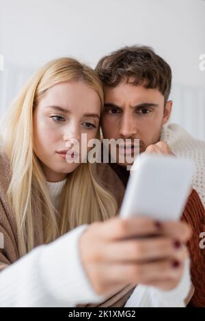 jeune femme blonde utilisant un smartphone près d'un petit ami inquiet couvert de couverture, image de stock Banque D'Images