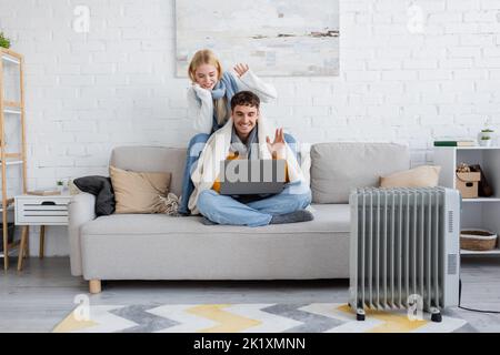 joyeux jeune couple dans les foulards et les sweaters en se vetant les mains pendant l'appel vidéo sur ordinateur portable, image de stock Banque D'Images