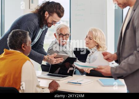 A young educator in a classroom with senior students shows lecture on tablet. Stock Photo