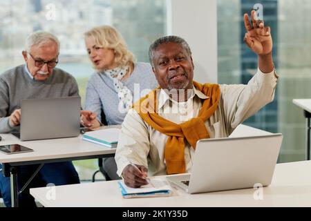 Seniors sit in a classroom and talking about project on the laptop while a multicultural senior student in foreground asking questions. Stock Photo