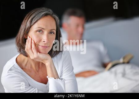 Commencez la journée dans un cadre détendu. Portrait d'une belle femme mûre assise sur son lit avec son mari en arrière-plan Banque D'Images