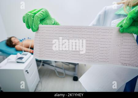 Test de cardiogramme du cœur de l'enfant gros plan dans les mains du médecin contre le fond du patient allongé sur un lit médical avec des sondes à vide dans la zone cardiaque Banque D'Images