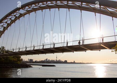 Le soleil du matin brille à travers le pont Humber Bay Arch pendant qu'une personne marche à travers. La tour CN et les gratte-ciel de Toronto sont vus en arrière-plan. Banque D'Images