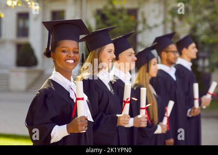 Une jeune femme afro-américaine étudiante en robe de remise des diplômes et un chapeau sourit avec les diplômés Banque D'Images