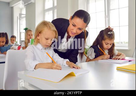 Jeune enseignante amicale aidant les petits élèves à effectuer des tests en classe primaire. Banque D'Images