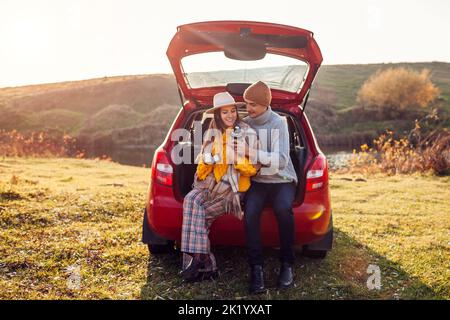 Un jeune couple amoureux se détend dans le coffre de la voiture au bord du lac d'automne en buvant le thé de la fiole à vide. Homme et femme voyagent en voiture. Les jeunes homme et femme apprécient l'automne Banque D'Images