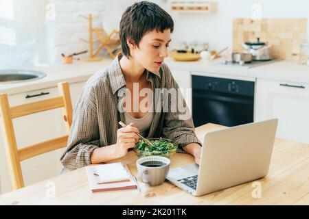 Une jeune femme occupée mange une salade de légumes dans la cuisine et travaille sur un ordinateur portable. Banque D'Images