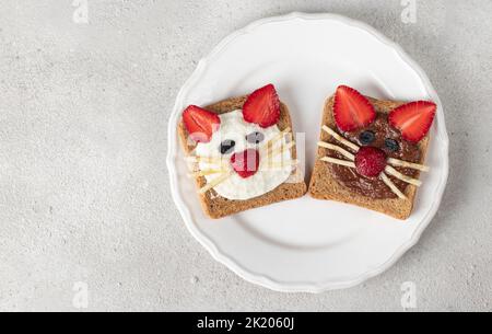 Deux toasts doux pour les enfants en forme de chat avec fraises, banane, fromage à la crème et chocolat sur plaque blanche sur fond gris, espace copie Banque D'Images