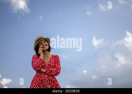 une jeune femme vêtue d'une robe rouge parle sur un téléphone portable sur fond de ciel bleu Banque D'Images