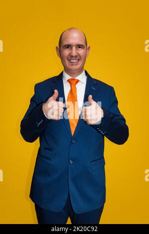 Portrait d'un homme cacaucasien souriant, vêtu d'un costume bleu, d'une chemise blanche et d'une cravate orange, les deux pouces regardant l'appareil photo. Le backgrou Banque D'Images