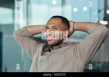 Gros plan d'un homme de course mixte qui se détend au bureau avec les mains derrière la tête et qui regarde par la fenêtre, souriant, homme d'affaires rêvant à l'intérieur d'un immeuble de bureau moderne, investisseur prospère content du travail Banque D'Images