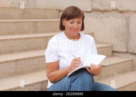 femme mature travaille à distance, l'enseignement supérieur pour les adultes, femme écrit et note dans un carnet assis sur les escaliers à l'extérieur, indépendant. Collège s Banque D'Images