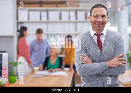 Le succès est une donnée avec moi en charge. Un beau jeune homme debout avec ses bras pliés pendant que ses collègues travaillent en arrière-plan Banque D'Images