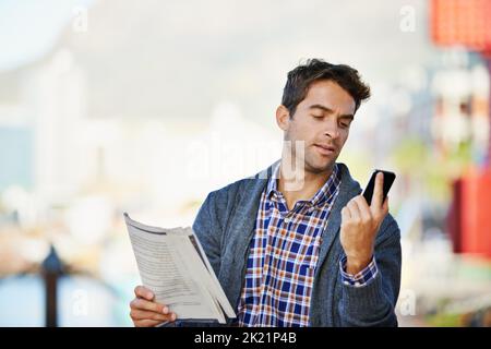 En comparant les nouvelles en ligne à l'ancienne, un beau homme tenant le journal et utilisant son téléphone à l'extérieur Banque D'Images