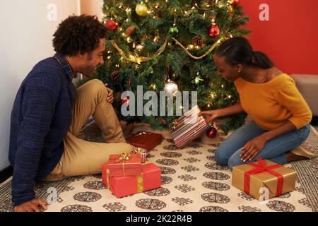 Tendre la main aux prézzies. Un jeune couple aimant assis avec leurs cadeaux à côté d'un arbre de Noël. Banque D'Images